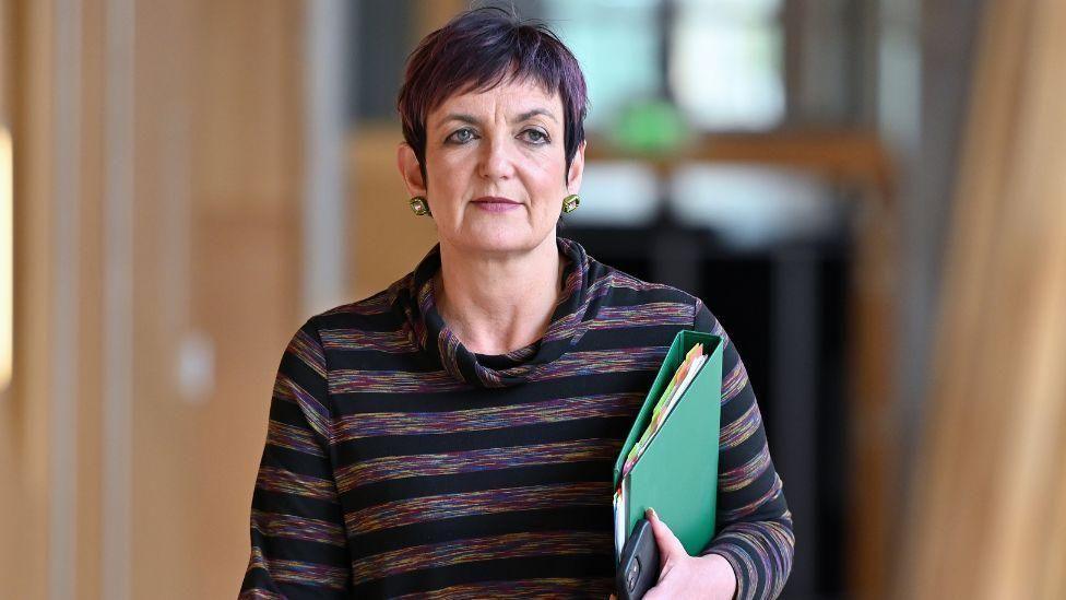 Angela Constance in a purple dress walks with a blue folder