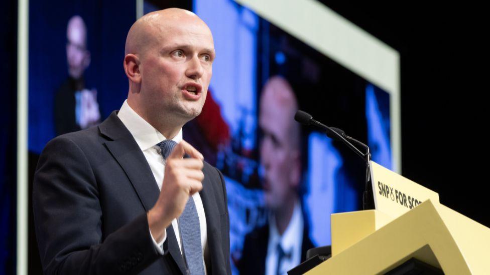 Stephen Flynn, a bald man in a dark blue suit with lighter blue tie, speaks at the podium of the SNP conference 