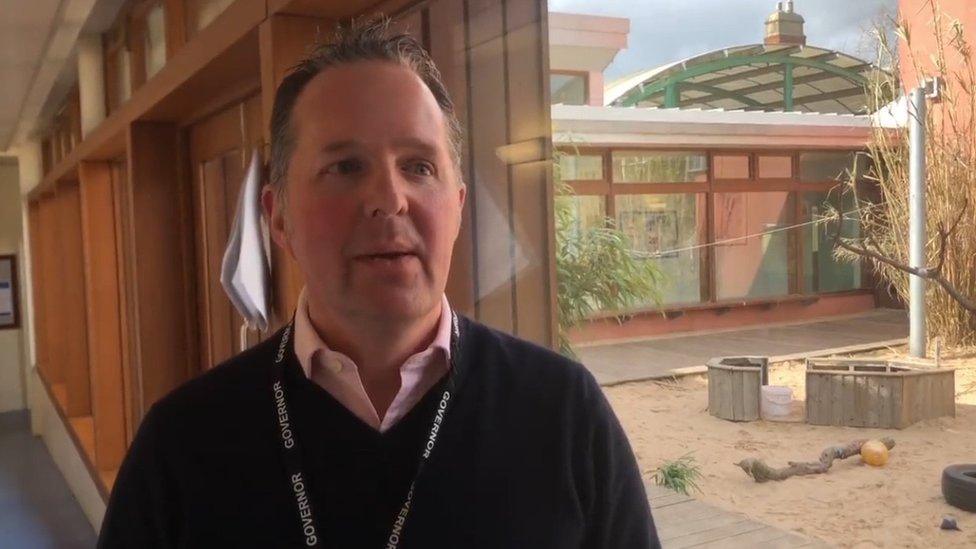 Man with short brown hair and wearing a lanyard stands in building that resembles a 1930s school