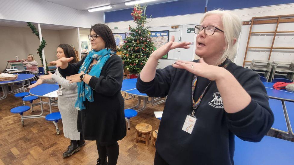 Three women in a school hall, standing in front of blue tables and benches, with a Christmas tree decorated in the background. They are making British Sign Language hand gestures.