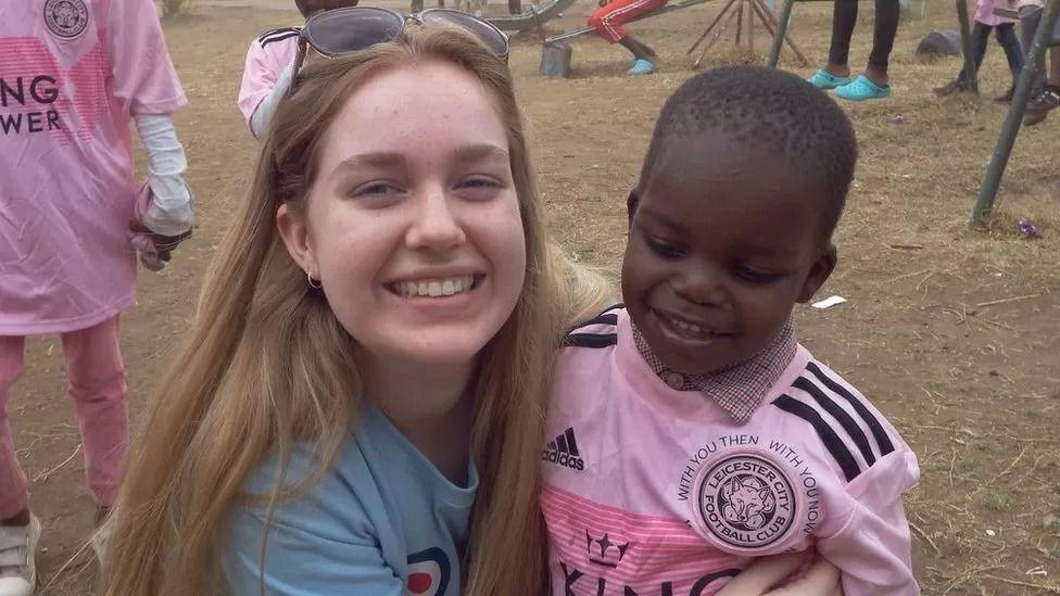 Charlotte Hope in Kenya with a young boy wearing a pink Leicester City Football Club shirt. Ms Hope is smiling and has sun glasses on her head