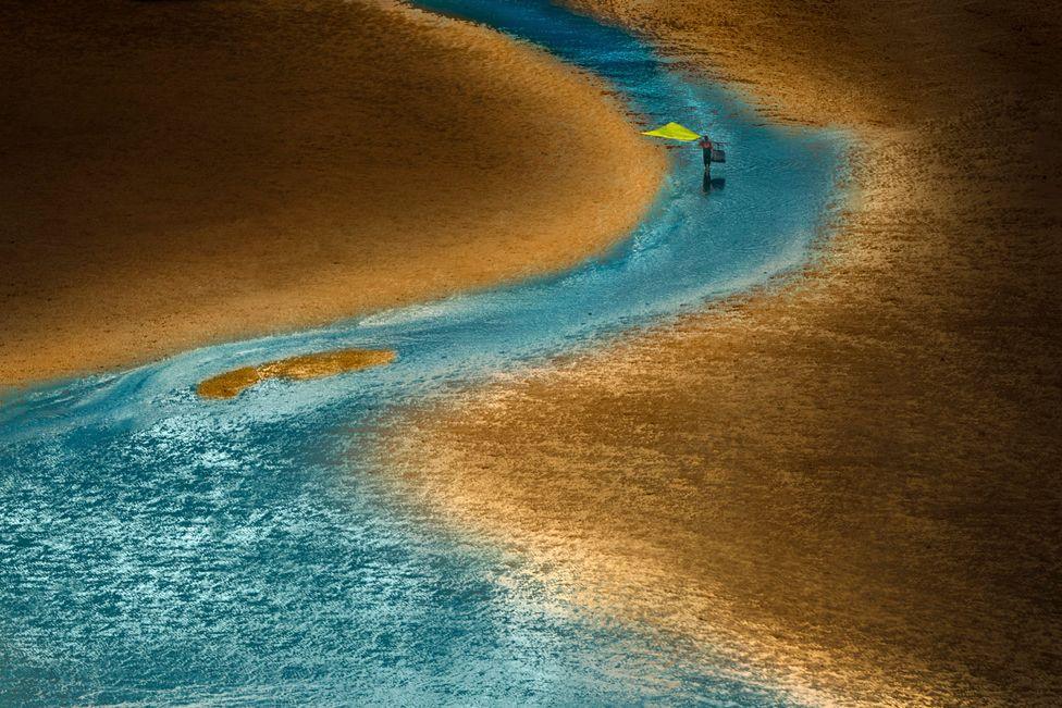 The golden haze of the sunset on a beach the while a traditional fisherman wades through the water in China
