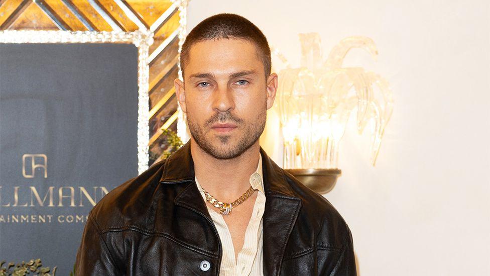 Joey Essex, a man wearing a black leather jacket and white shirt, with a chain around his neck. Behind him is a lamp and a promotional brand board.