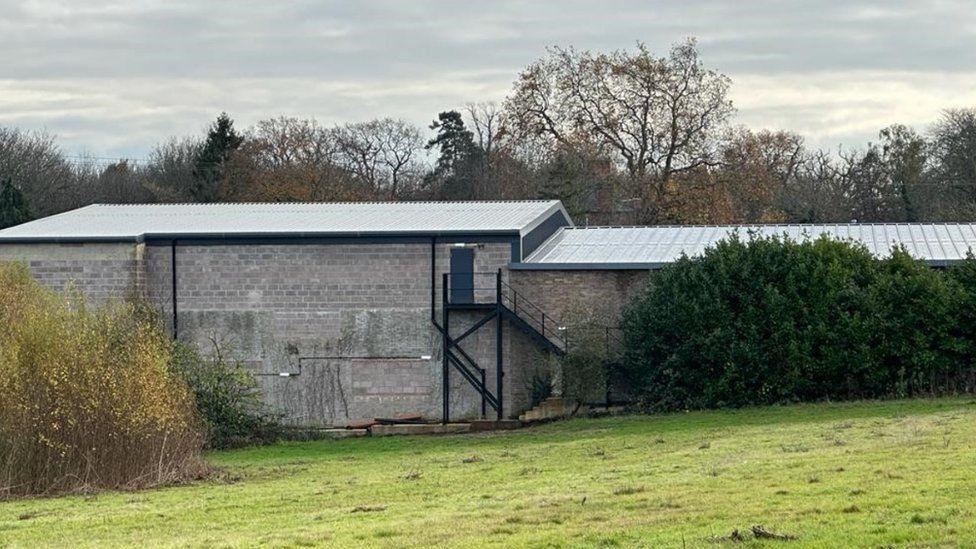 Rear of grey brick shed-like building with external stairs up to a top floor door and field in front