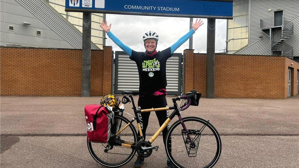 Kate Strong standing outside the football stadium alongside her bike with her arms in the air