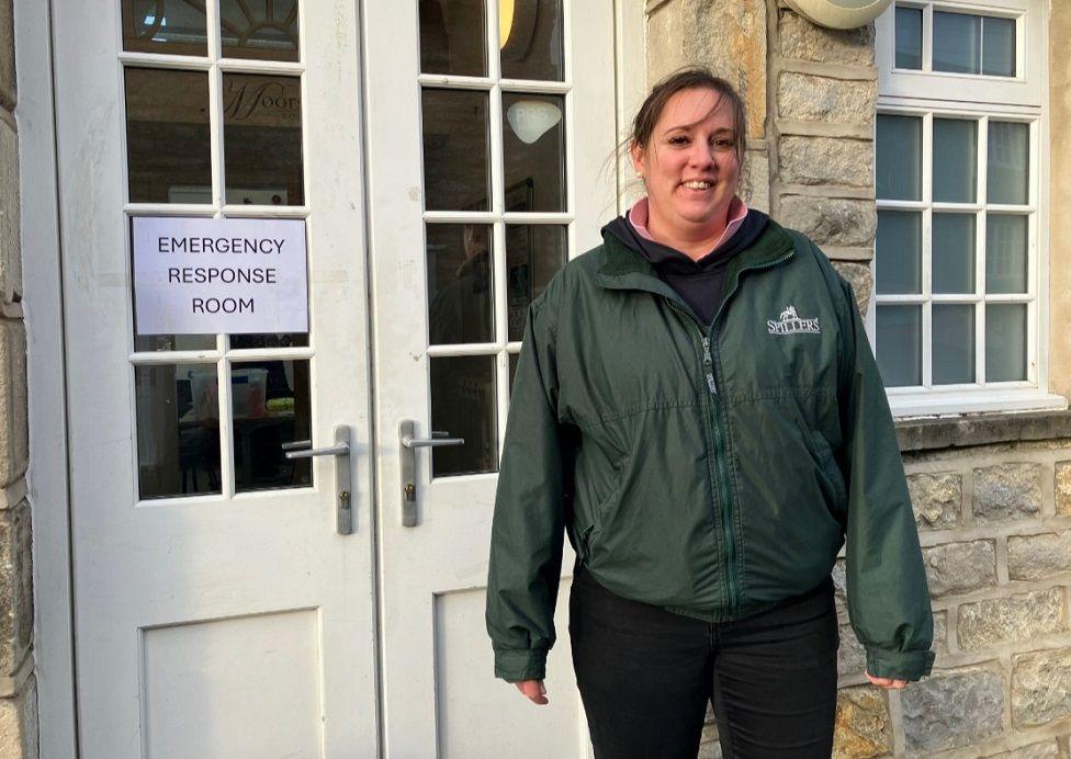 Lisa Young stands in front of a white door with a sign saying 'Emergency Resposne Room'. She is wearing a green rain coat and black jeans 