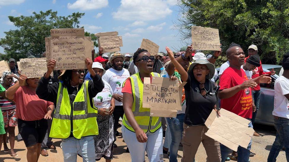 A crowd of people wave hand-drawn placards during a protest at the mine