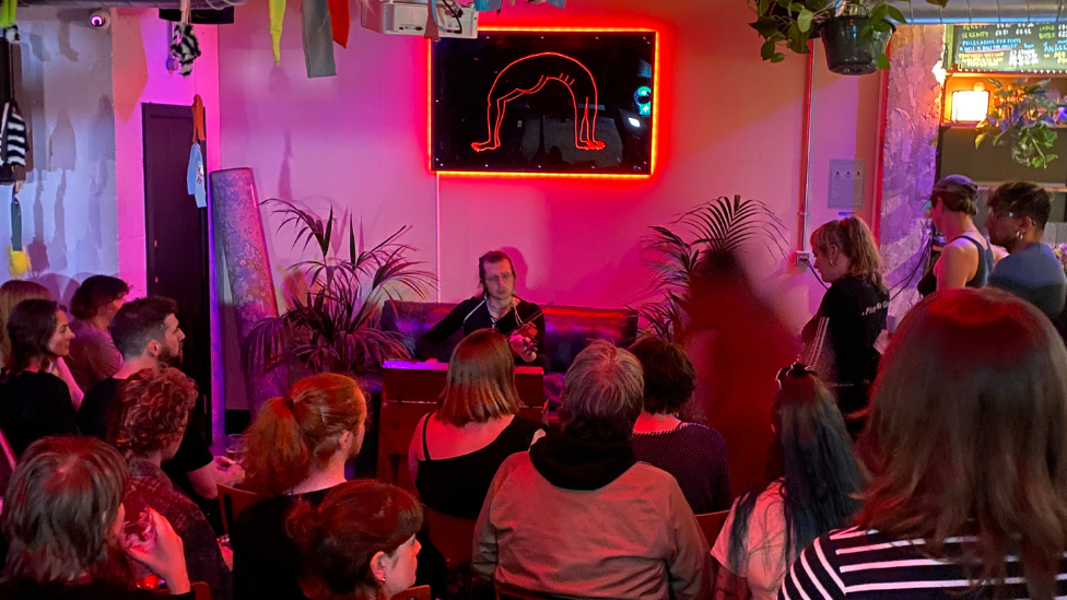 A crowd watch a violinist who is playing while sitting on a sofa. The lighting is softly pink