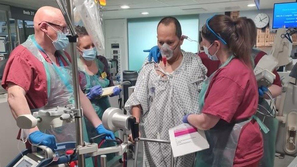 Mr Franco being supported by a team of medics in scrubs, while he walks with a frame and is hooked up to an oxygen mask.