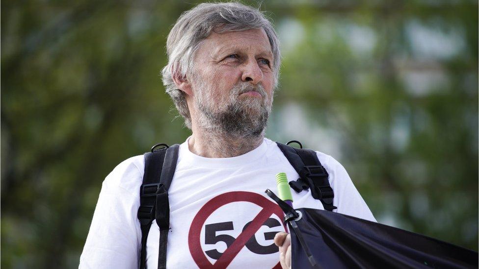 A man wearing a white t-shirt with a 5G symbol and a red cross through it holds an obscured banner at a protest