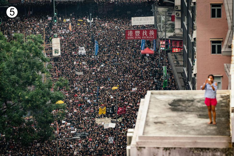 A woman stands on a rooftop taking a photo of the crowd below in this artistic composition