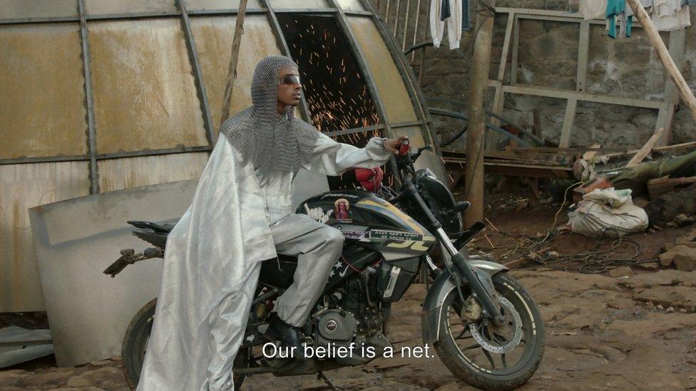 A still from "Except this time nothing returns from the ashes", showing a character sat on a motorbike. The caption reads "our belief is a net"