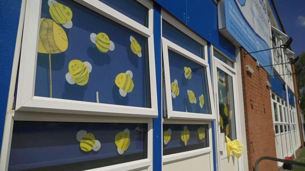 Cardboard cut outs of yellow bees are displayed windows with white window frames and blue blinds behind.