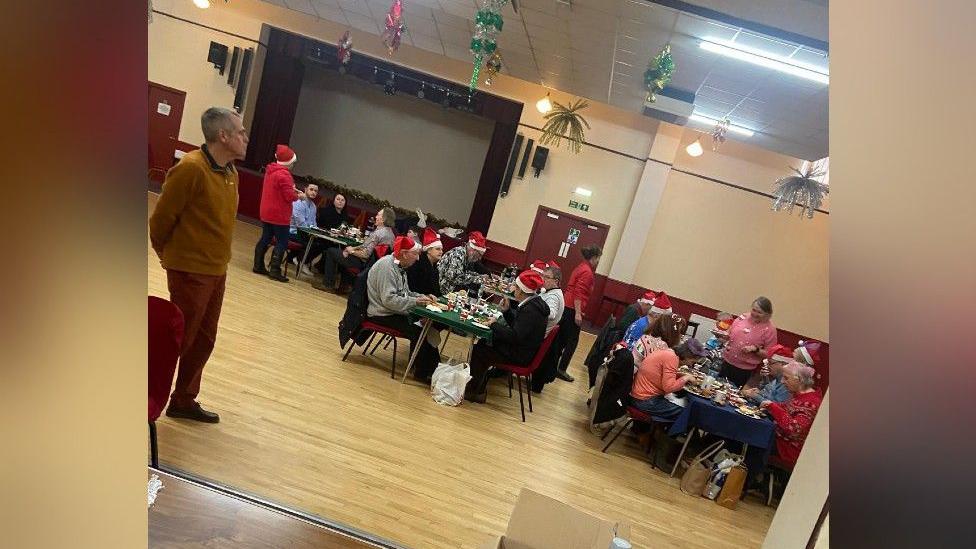 Tables in a hall have people sitting at them, eating dinners. Many of the people are wearing red and white santa hats. There are christmas decorations hanging from the ceiling.