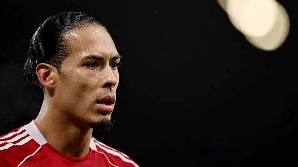 Close-up image of Virgil van Dijk in the left of the photograph. He has black hair, tied back, and is wearing Liverpool's red home shirt. The background of the image is black with a spotlight in the top right corner.