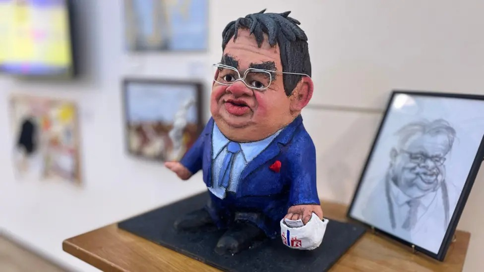 A papier-mâché caricature of Guernsey deputy John Gollop, with similarly grotesque pencil sketch behind. He is wearing an ill-fitting blue suit with his shirt untucked. He wears glasses and holds a plastic carrier bag stuffed with papers in one hand and a lit cigarette in the other.