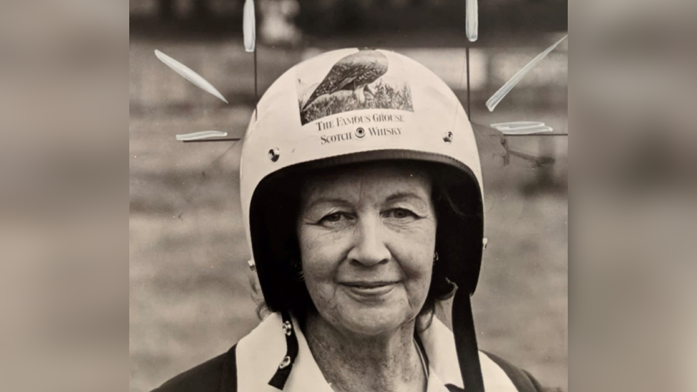 A black and white photo of Hugo's grandmother, a middle-aged woman wearing a crash helmet with "the famous grouse scotch whisky" on it