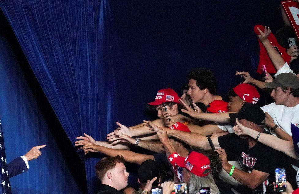 A group of people on the right, some wearing read baseball caps, stretch their hands forward to touch the hand of Donald Trump as he points out from the left behind a flag in front of a blue curtain