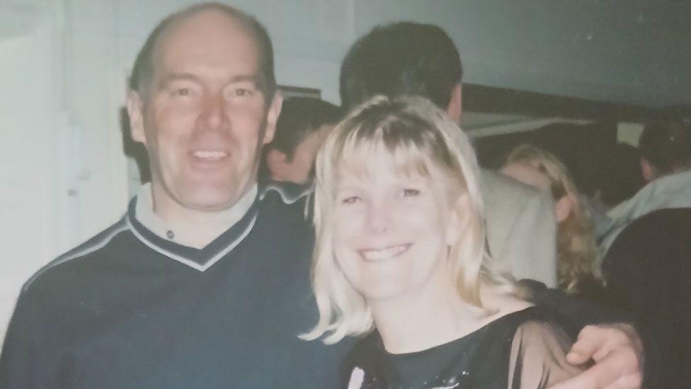 A faded colour photo of Alan Marshall on the left with his arm around his sister Janet Newell. He is wearing a blue jumper over a white shirt and is clean-shaven. She has shoulder-length blonde hair and is wearing a black dress. Both are smiling broadly. They are in a large room and there are people chatting behind them. 