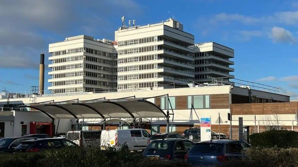 External view of the main Lister Hospital building, opened in 1972. An incinerator chimney and the red coloured entrance to the Emergency Department and Urgent Care Centre can be seen to the left. Ten vehicles are parked outside.