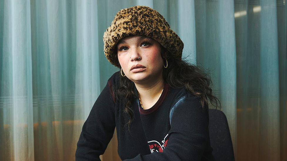 Lola Young wearing a black long sleeved top, looking into the camera, wearing a printed winter hat, with light blue coloured curtains behind her.