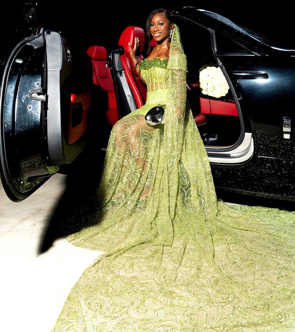 Nian Fisher sitting by the open door of a black limousine. Her green dress has a train that is arranged in front of her.