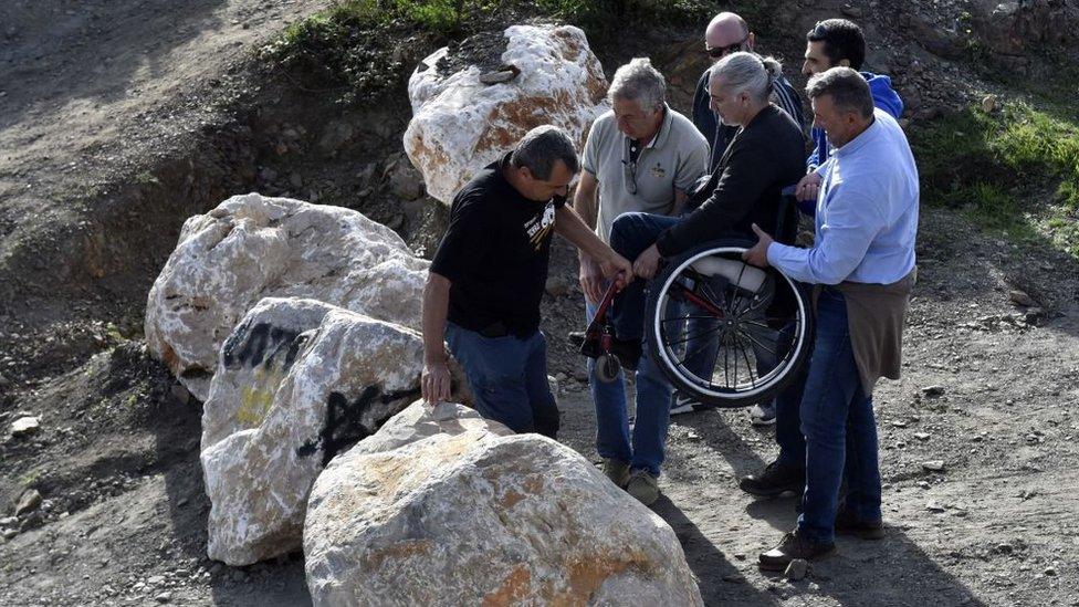 People lift a woman using a wheelchair over some rocks blocking a road over the Franco-Spanish border