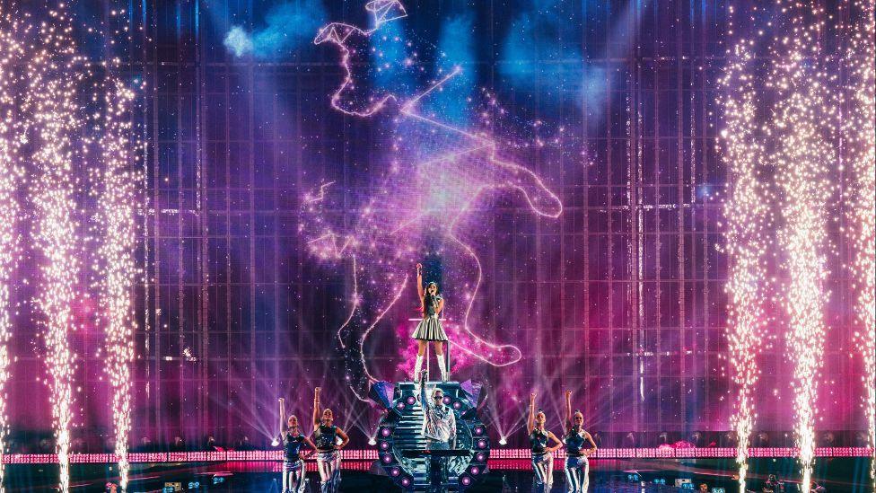 A stage setting with a performer in a silver dress raised on a podium, there are lots of pyrotechnic sparks either side, with two dancers on either side.
The outline of a dog, styled like a star constellation, is projected on the wall behind with purple lighting