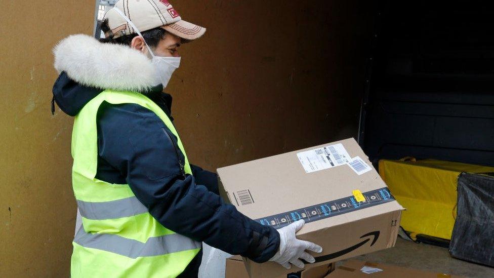 A delivery man wearing a protective mask carries an Amazon package.