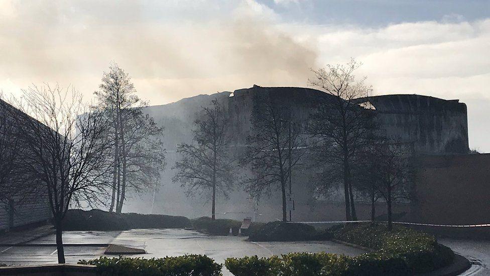 A large building destroyed by fire with smoke surrounding the premises. There are trees in front and police tape around the building.