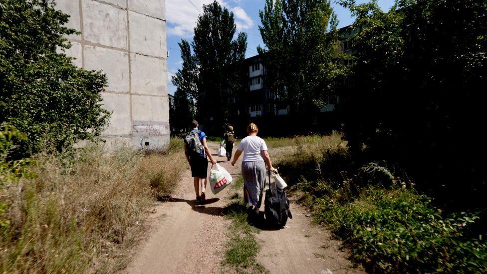 Three people walk down a dirt path past a building and piles of weeds, carrying large bags
