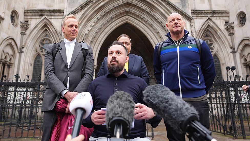 Martin Hibbert (centre), who was injured in the May 2017 Manchester Arena bombing, makes a statement outside the Royal Courts Of Justice, central London in July. Three people , including his lawyer, stand behind him