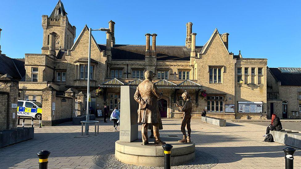 Lincoln Central Station a two-storey of light stone with pitched roofs, chimneys and gothic tower. In the foreground is a statue of Victorian mathematician George Boole, depicting Boole lecturing two children