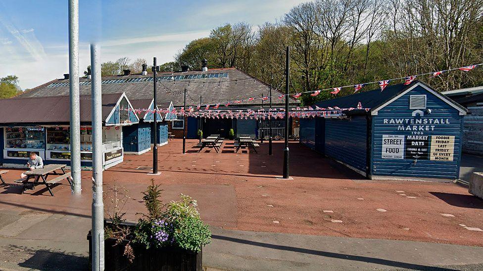 Street view of the current market, with three single storey open stalls with pointed roofs to the left, a large building in the middle and a blue wood building to the right. Union flag bunting is strung between the buildings, which sit on a wide, red asphalt floor