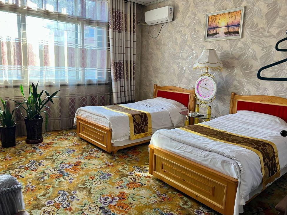 A hotel room with two beds, yellow floral carpet, potted plants, a framed landscape picture and windows with decorative blinds