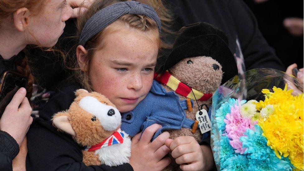 Girl holding corgi and Paddington Bear toys waiting for funeral procession to pass.