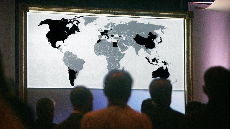 A map of the world shown on a screen with people standing in front of it silhouetted.