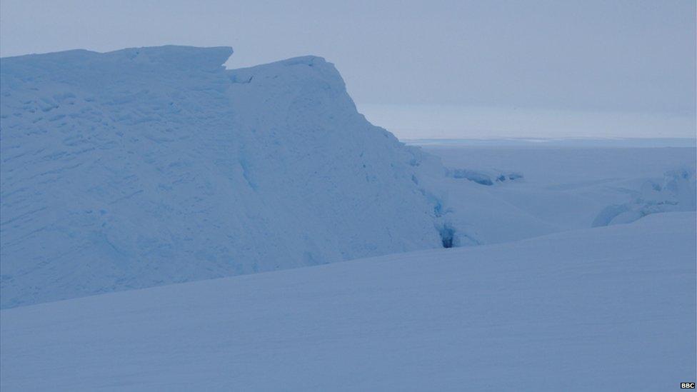A landscape of pure white snow, with a few dips in the surface and a big mound on the left hand side. The sky is white.