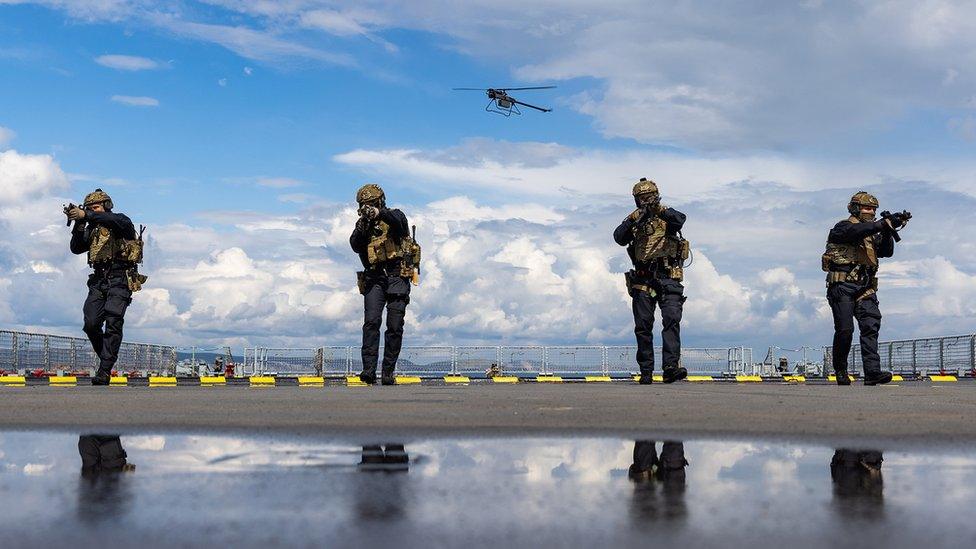 Four marines take combat stances with a drone flying overhead