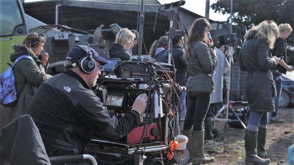 A film crew and camera man sat behind a large camera and filming equipment. There are lots of people standing around in coats and wellies on a muddy patch of ground, wearing headphones and facing in the direction of the actors.