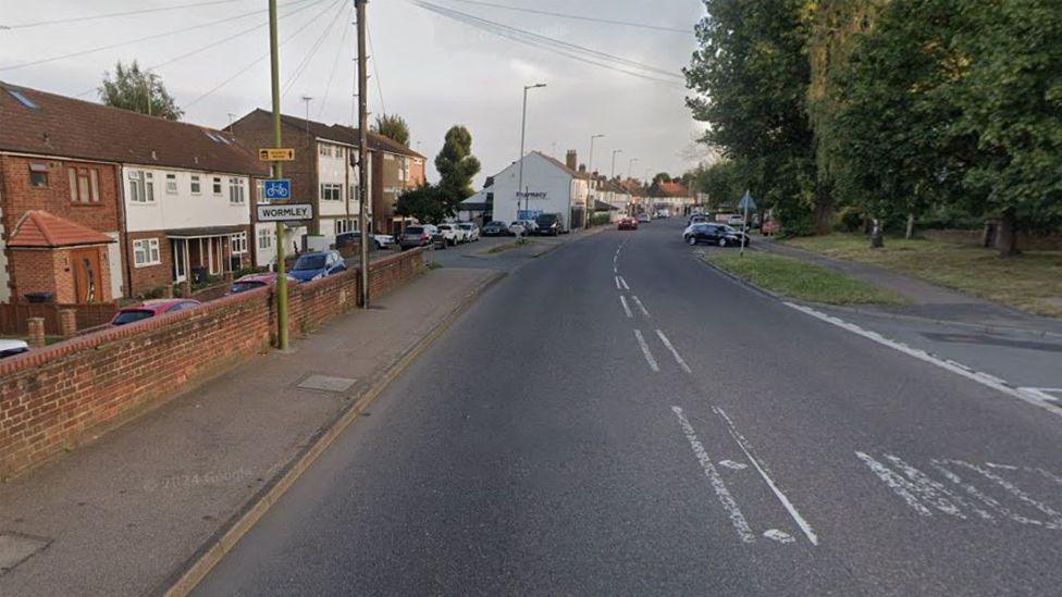 Wormley High Road at one end. There is a pavement on the left bordered by a low brick wall and terraced houses behind it which continue into the distance. The right side of the road has a grass verge, pavement behind it and some green trees. 