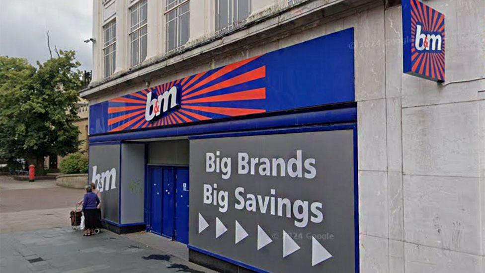 The outside of B&M in Bedford, with two women chatting outside the store. Above the store front is a big bright B&M sign in blue and orange and a vinyl wrap saying "big brands big savings" in a large window.