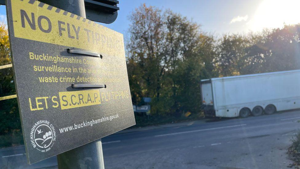 A black sign saying No Fly Tipping on black letters on a yellow rectangle. Other lettering in yellow says the site is under surveillance by Buckinghamshire Council. It is on Mumford Lane opposite a lay-by where the rear end of a white dumped articulated lorry trailer can be seen