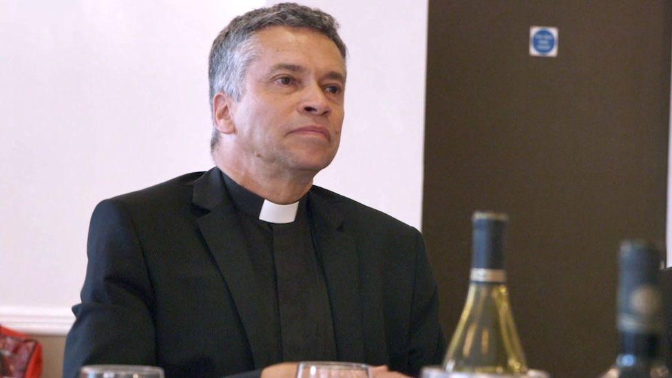 David Tudor, a former vicar in Essex, pictured taking part in a BBC documentary in 2018. He sits at a table, wearing the traditional Church of England attire of a black suit with a white dog collar, with greying hair. There are wine bottles and glasses on the table in front of him, suggesting the meeting he is attending is somewhat informal.