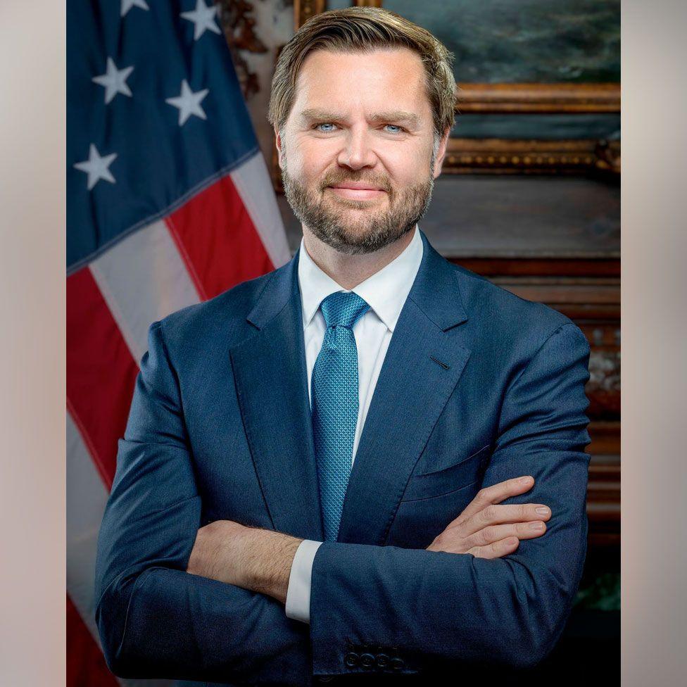 JD Vance stands with his arms crossed and a slight smile, wearing a blue suit and tie.