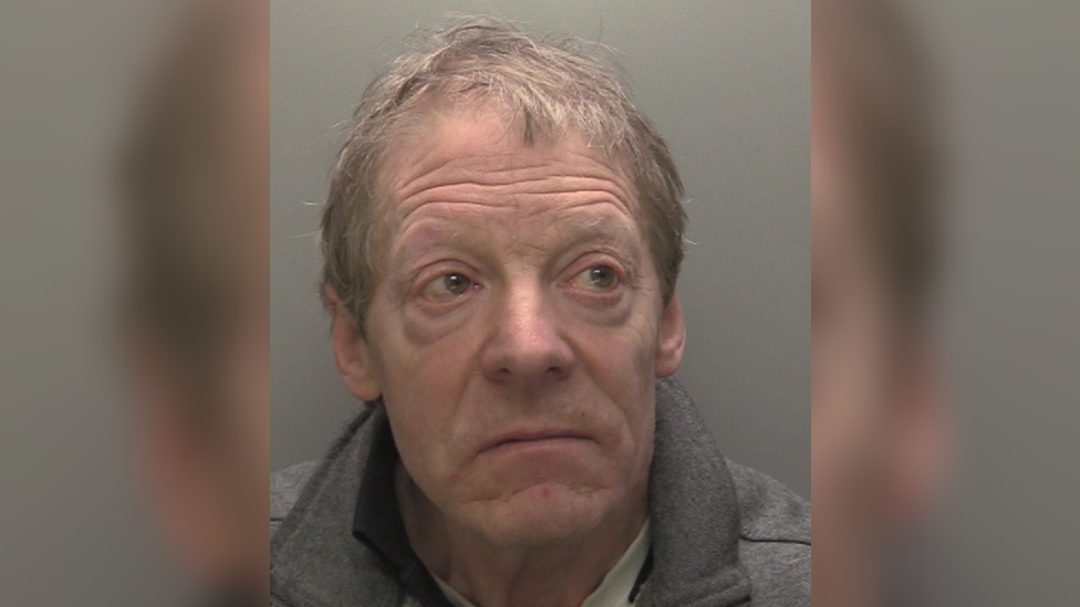 A police custody shot of David Glover. He has short grey hair and is wearing a grey collared jumper. He is looking to the right of the camera. The wall behind him is grey.