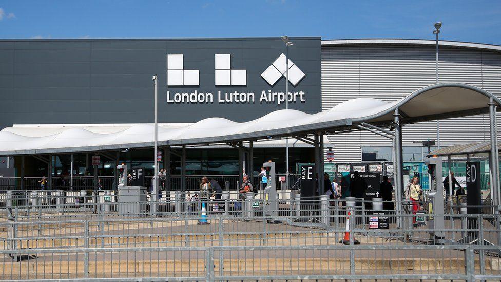 An entrance to London Luton Airport. It is a grey building with London Luton Airport written in white on its wall. In front of it are barriers and a white covered walkway. People can be seen walking along