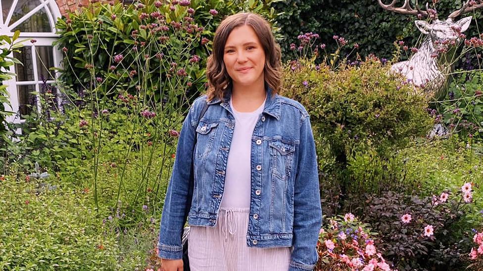 Sophie, wearing a light top with a denim jacket, with greenery and flowers behind her.