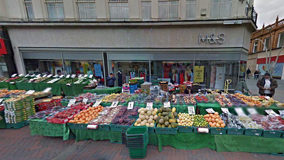 The outside of the former M&S, showing its scaled back logo above the door. It is market day, and outside the front of the store is a big fruit and veg stall.