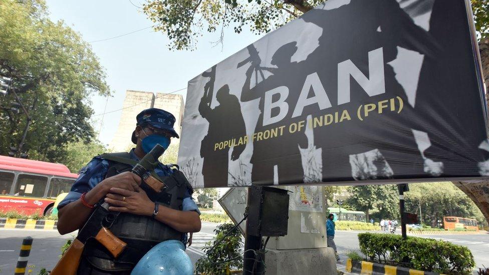 A big poster demanding a ban on the PFI next to a security officer holding a gun and wearing body armour